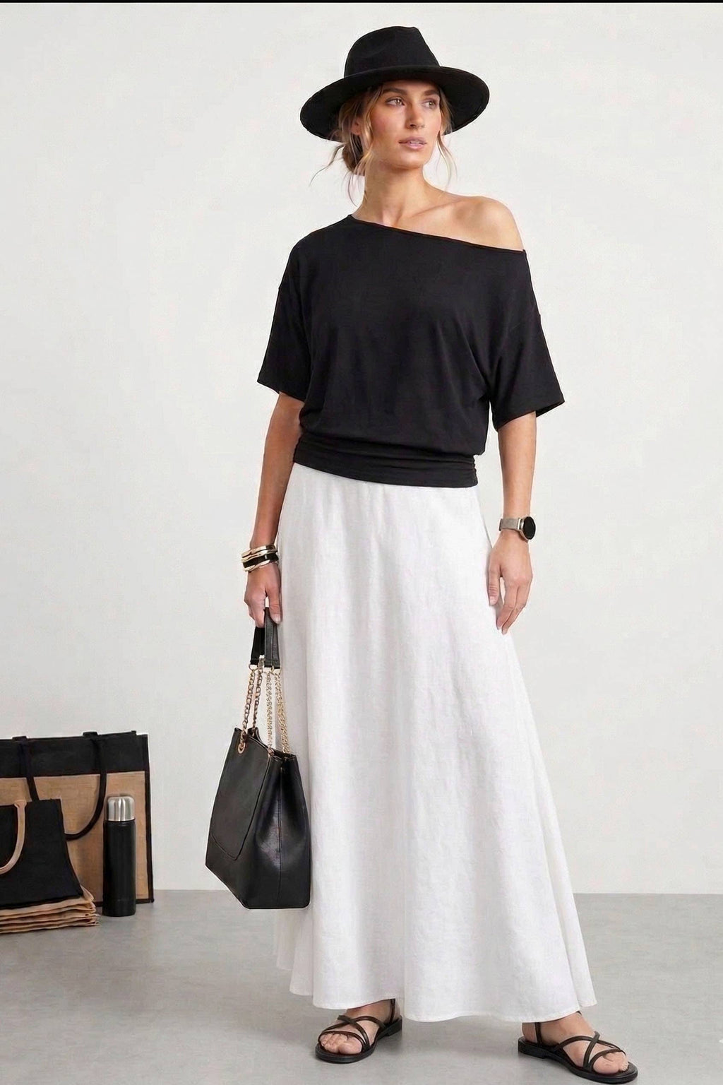 Woman wearing a black off-shoulder top and white pants with a black hat and bag on a white background