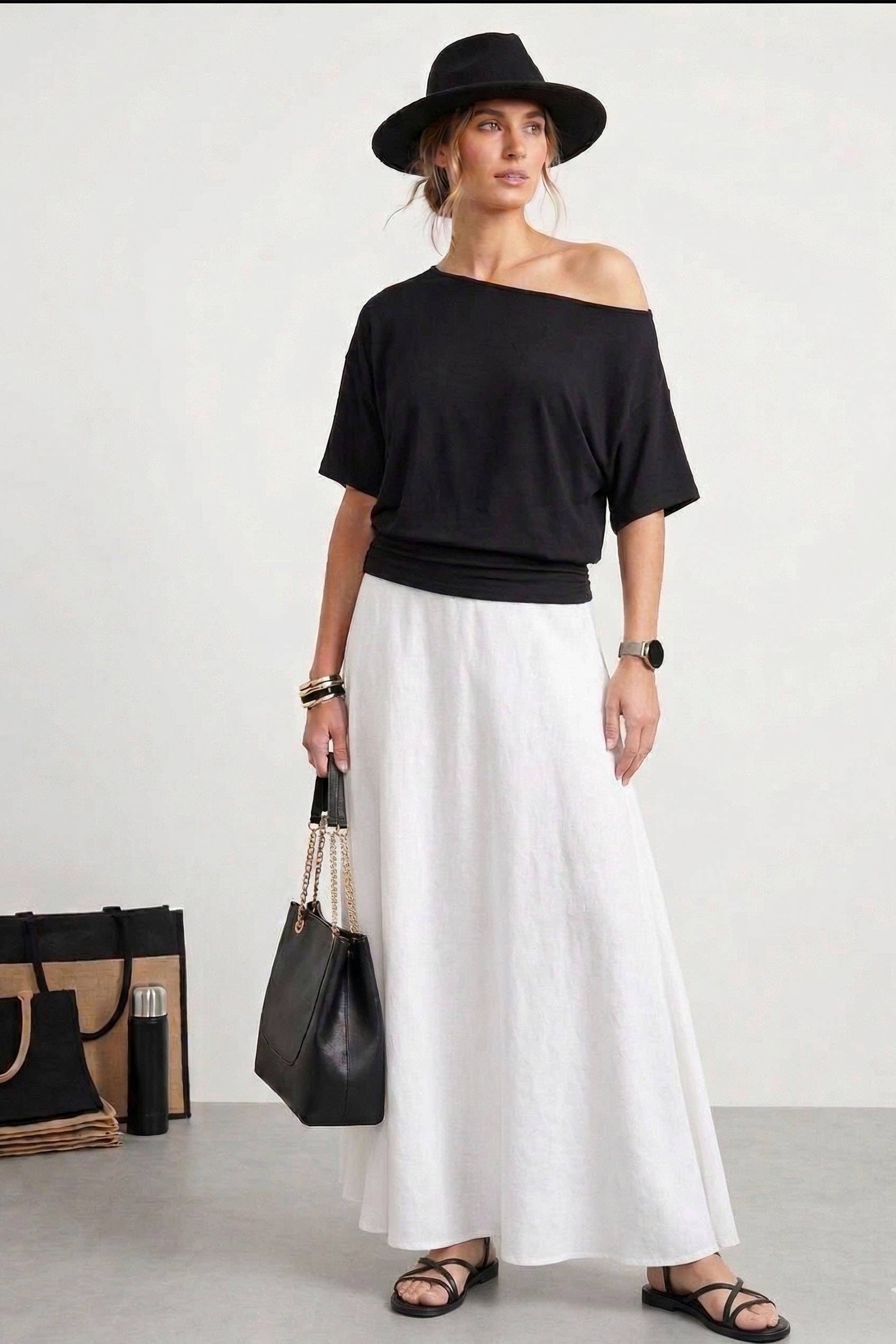 Woman wearing a black off-shoulder top and white pants with a black hat and bag on a white background