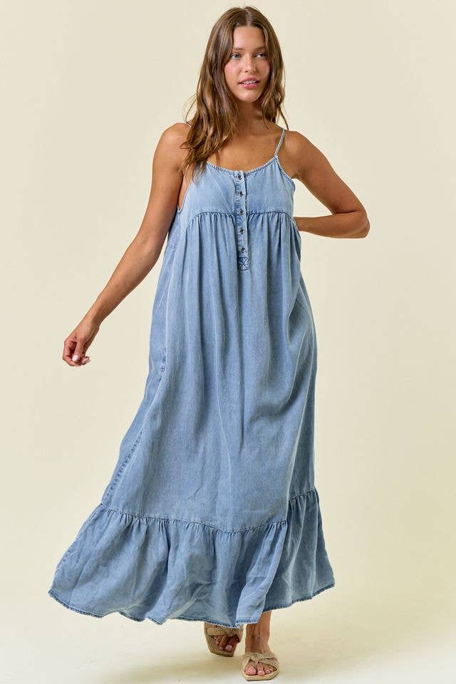 A woman modeling a light wash denim chambray maxi dress featuring adjustable spaghetti straps, a partial button-down front bodice, an empire waist, and a ruffled tiered hemline.
