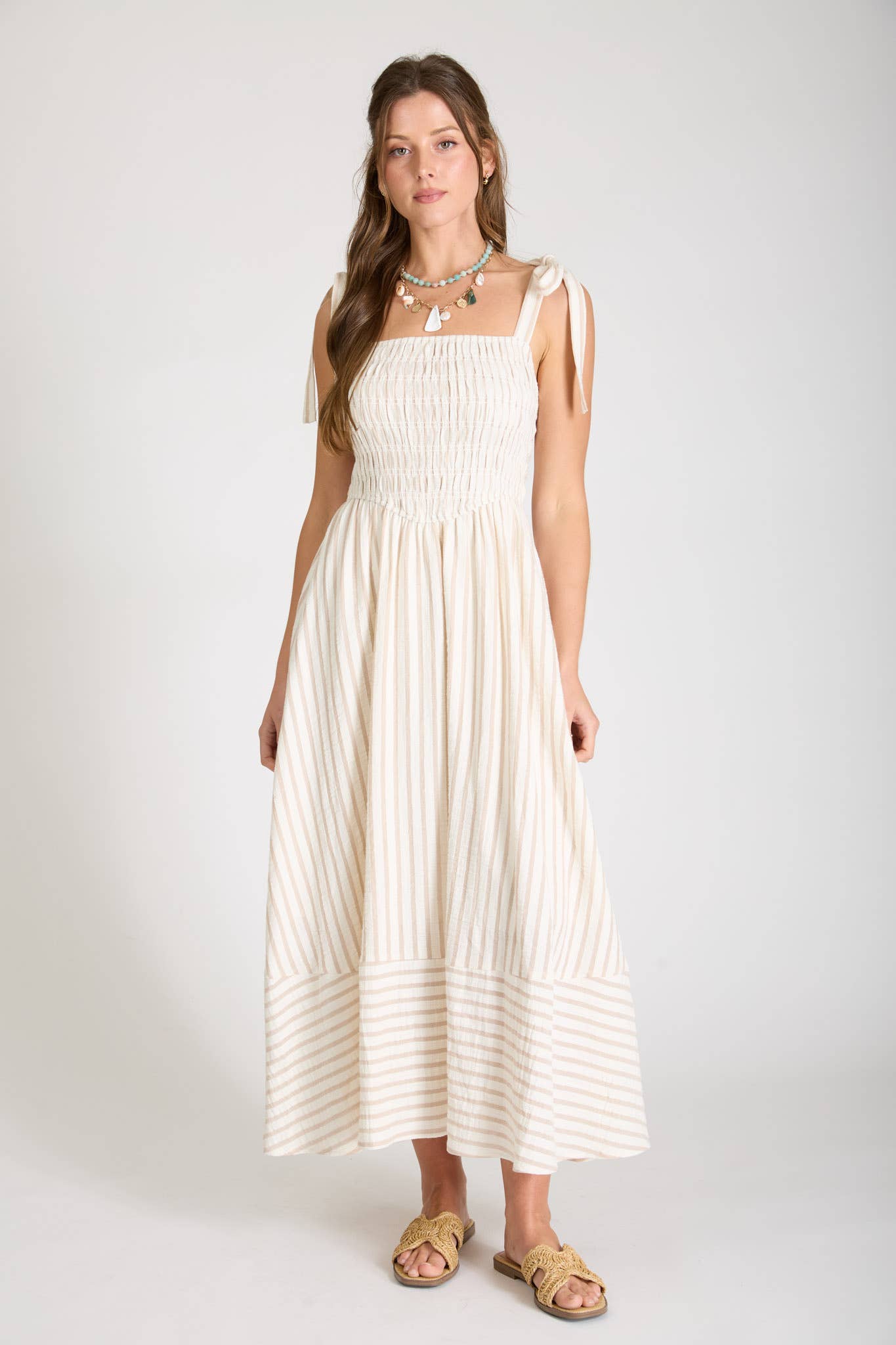 A woman posing against a white background wearing a cream and tan vertically striped midi dress, featuring a long smocked bodice, tie-strap shoulders, and a matching horizontal stripe pattern along the hemline on  a plain background