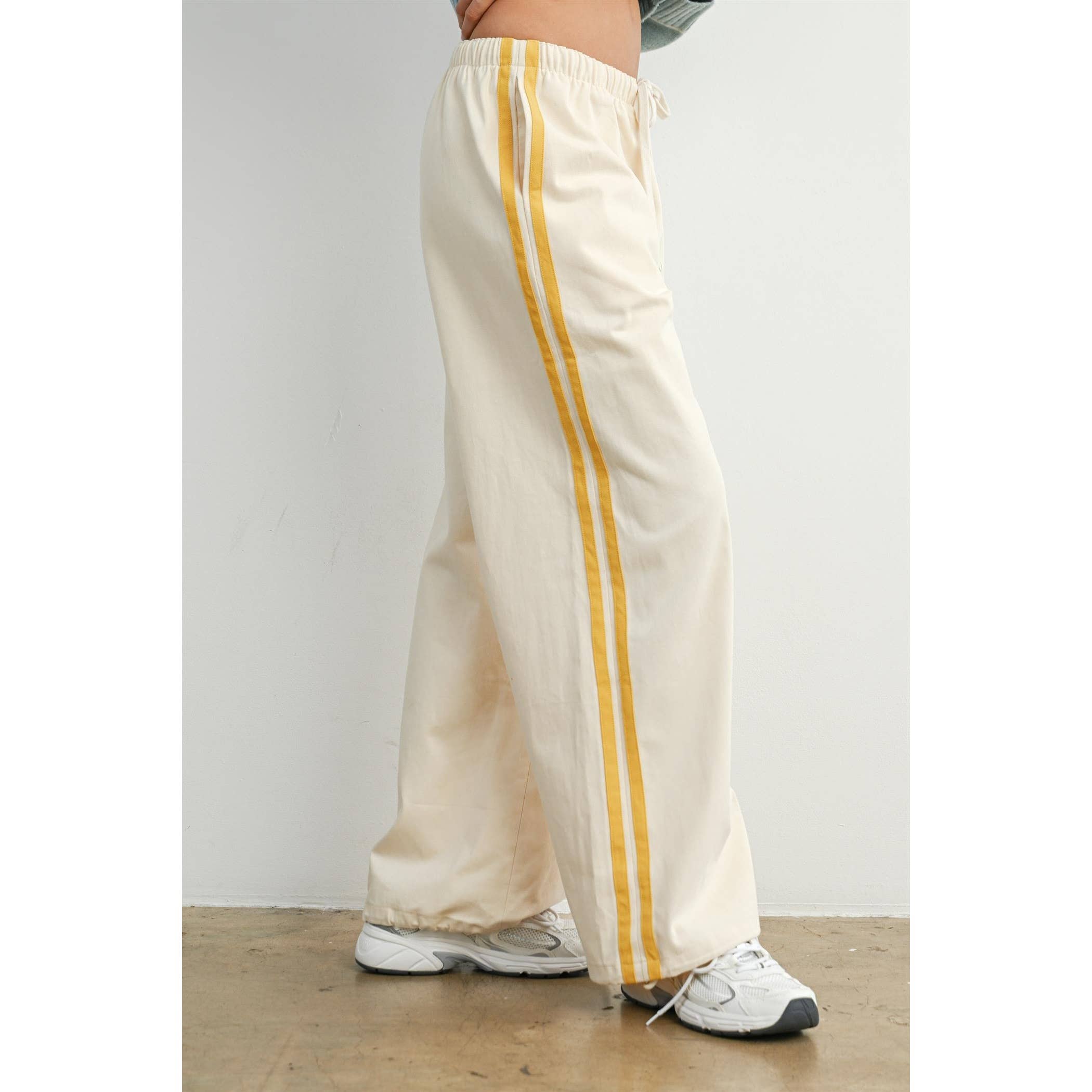 A close-up side view from image_2.png showing a model wearing relaxed-fit off-white trousers with a prominent yellow side stripe and sporty toggle drawstring cuffs at the hem worn by a person on a plain background