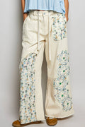 Off-white wide-leg utility pants featuring eclectic patchwork details including blue floral prints, white lace, and large embroidered flower patches. The pants include a drawstring waist and raw-edge hems for a bohemian, distressed look on a plain background 