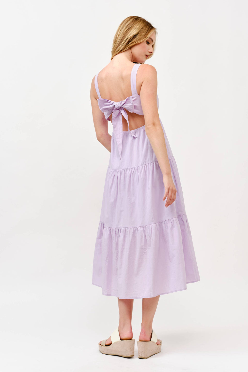 Rear view of a woman wearing a light lavender midi-length sun dress. The dress features wide tank straps, a large decorative bow tie at the mid-back with a cut-out detail, and a three-tiered flowy skirt. She is wearing cream-colored platform wedge sandals against a white background.