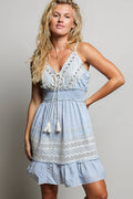 A woman wearing a light blue sleeveless mini dress with white eyelet floral embroidery on the bodice and skirt. The dress features a V-neckline with a rope lace-up tassel detail, a stretchy smocked waistband, and a ruffled hemline on a gray background