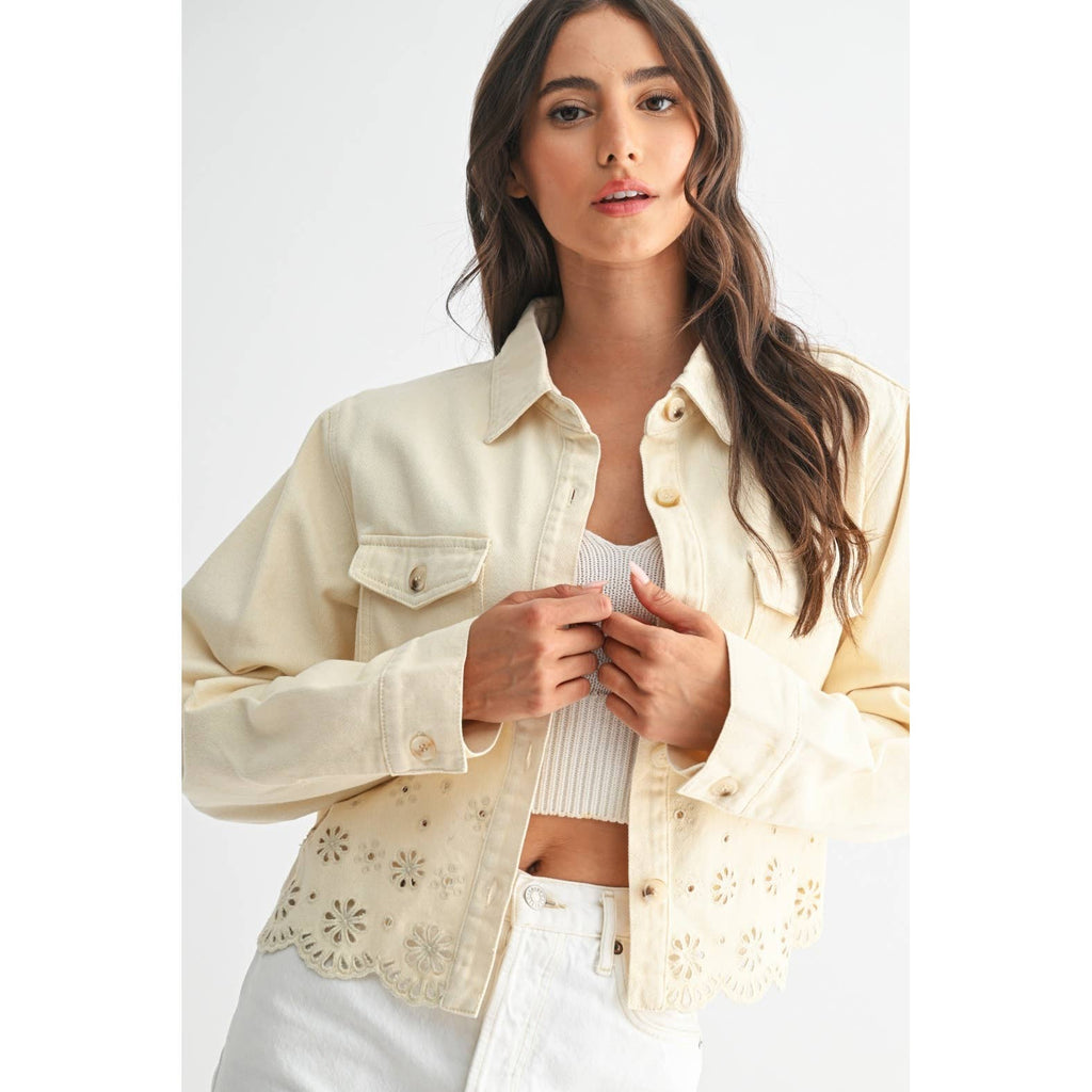 A woman wearing a cream-colored, long-sleeved cropped denim jacket featuring a scalloped eyelet lace hem with floral embroidery. The jacket has a collar, button-down front, and chest flap pockets, styled over a ribbed white crop top and white jeans on a plain background 