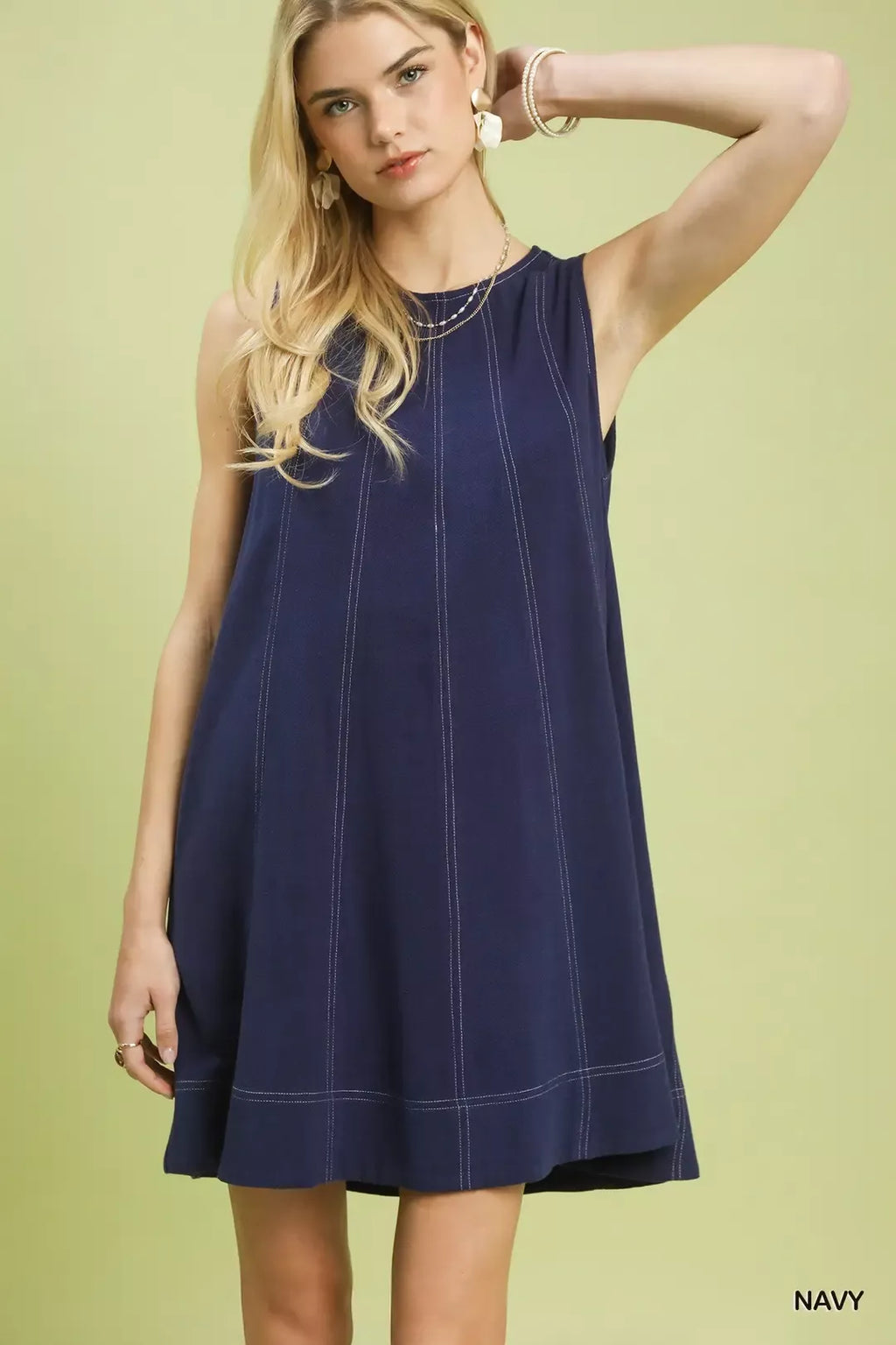 Model wearing a sleeveless navy blue shift dress with white contrast vertical stitching and a flared hem against a light olive background.