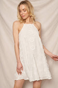 Ivory sleeveless swing dress featuring delicate floral embroidery, a high halter neckline, adjustable spaghetti straps, and a flowy A-line silhouette with textured floral details throughout against a beige curtain background