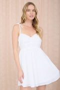 Woman wearing a spaghetti strap mini dress with adjustable tie straps, lace-trim sweetheart neckline, button-front detail, and a flowy A-line skirt against a beige background