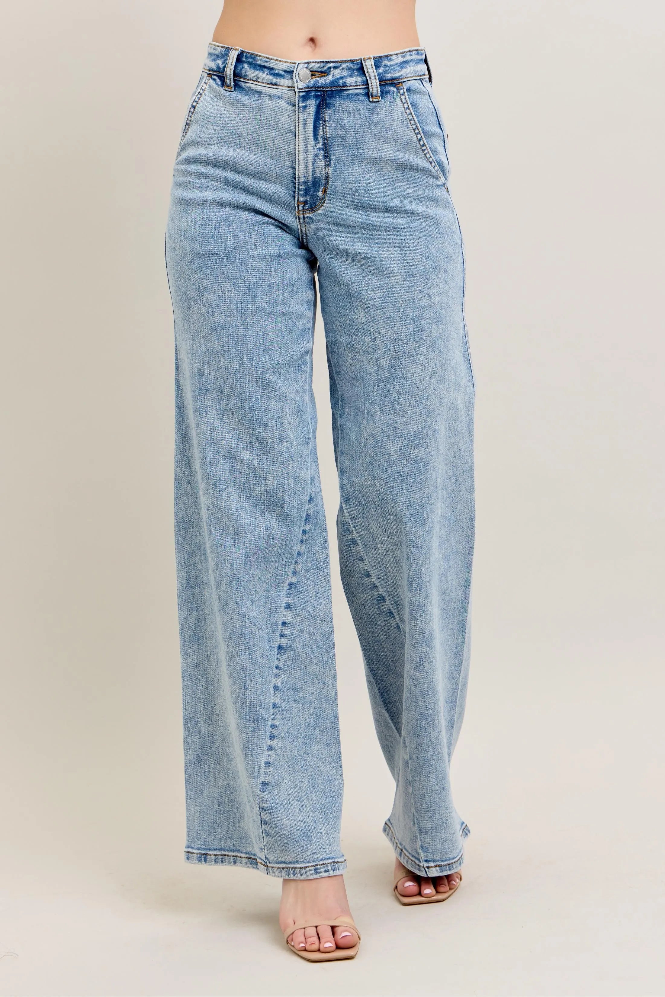 Front view of a person wearing high-waisted, light-wash wide-leg denim jeans with unique diagonal seam detailing and a relaxed fit on a beige background.