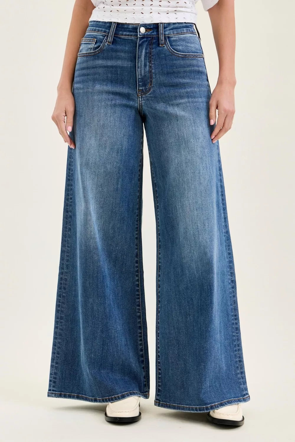 Front view of medium-wash, high-waisted wide-leg jeans with a floor-length hem, classic five-pocket styling, and subtle whiskering at the hips with white loafer shoes on a plain background 