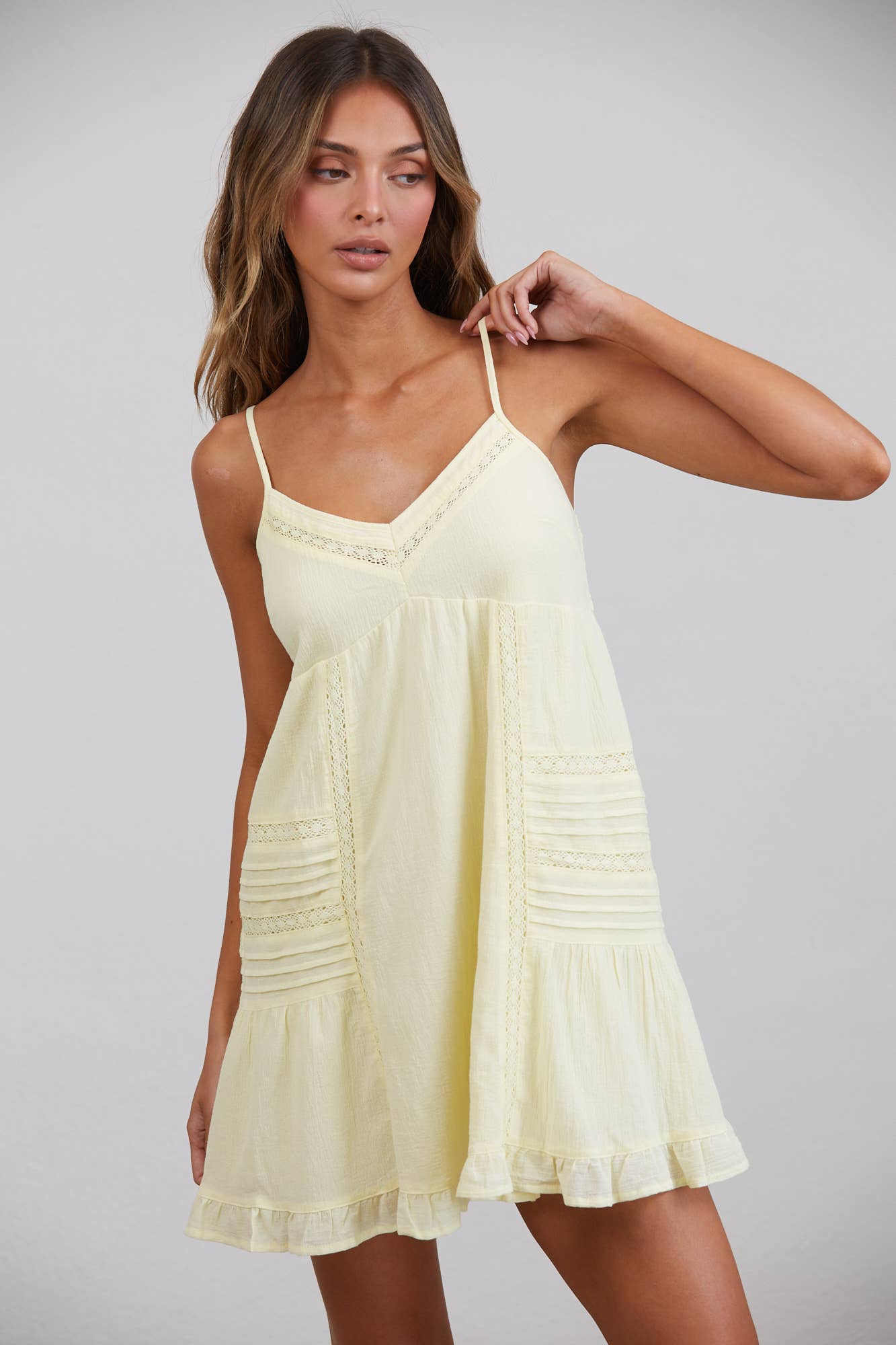 A front view of a model wearing a light pastel yellow sleeveless mini dress. The dress features thin adjustable spaghetti straps, a V-neckline trimmed with delicate white lace, vertical crochet panels, horizontal pintuck pleating on the sides, and a flirty ruffled hemline against a plain background