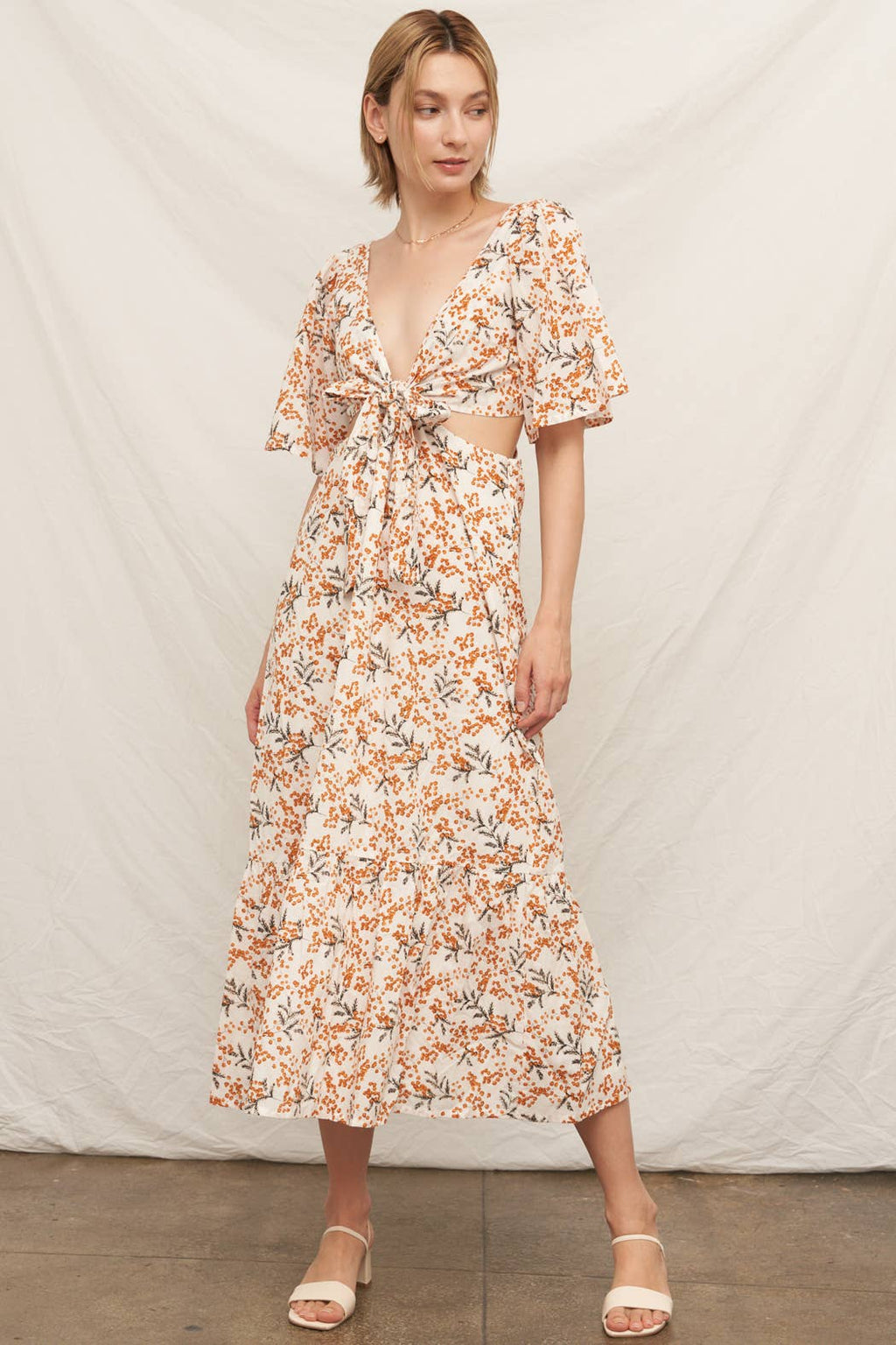 A woman posing in a cream-colored maxi dress featuring an all-over orange and brown floral print. The dress has short flutter sleeves, a deep V-neckline with a tie-front bodice, side waist cutouts, and a tiered ruffle skirt with a white background