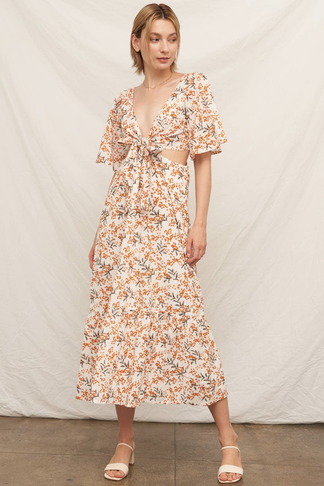 A woman posing in a cream-colored maxi dress featuring an all-over orange and brown floral print. The dress has short flutter sleeves, a deep V-neckline with a tie-front bodice, side waist cutouts, and a tiered ruffle skirt with a white background