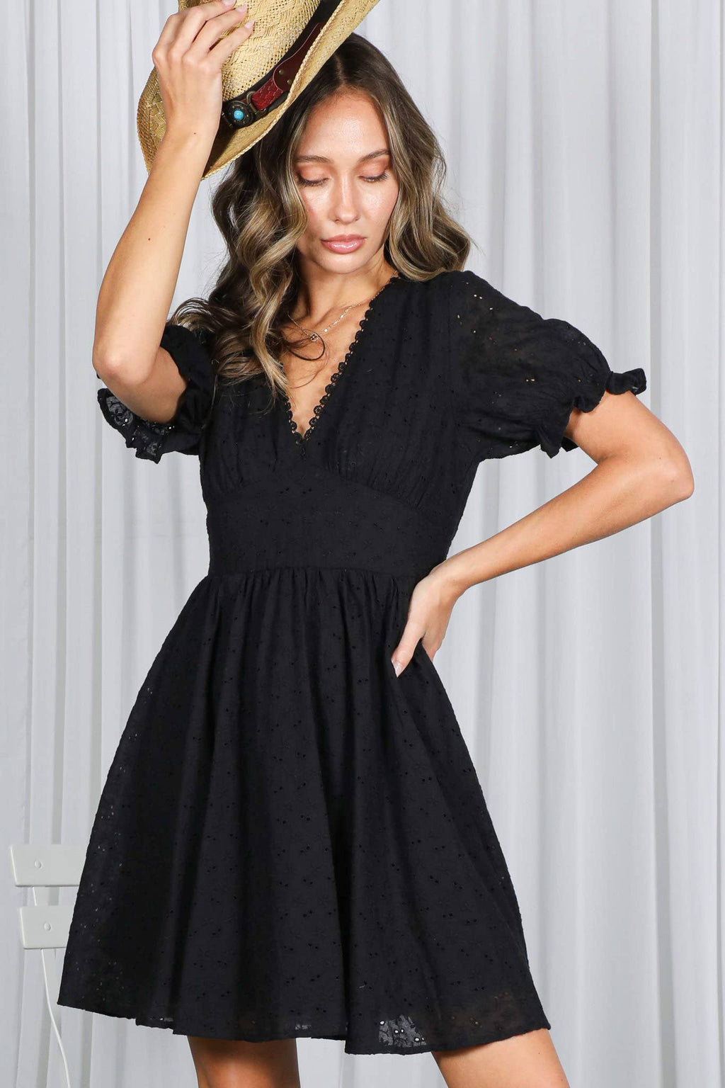 A woman wearing a black eyelet mini dress featuring a deep V-neckline with delicate lace trim, puff sleeves with ruffled cuffs, and a defined imperial waist. A model in image.png poses with a hand on her hip, tipping a straw cowboy hat against a white draped curtain backdrop 