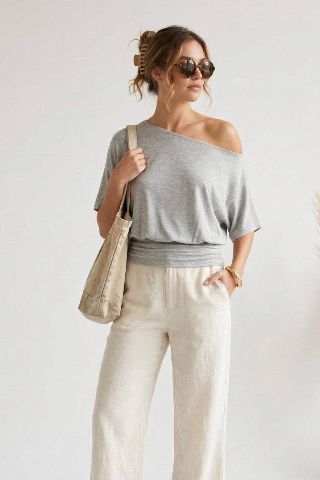 Studio shot from image_7.png of a model wearing an asymmetrical heather grey dolman tee styled off one shoulder with a thick banded hem, accessorized with round sunglasses and a canvas tote bag in a white background.
