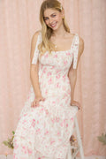 A blonde woman smiling while wearing a white maxi dress with a pink floral print, featuring a smocked bodice, tie-strap shoulders, and a tiered ruffle skirt with swiss dot fabric texture standing on a stool against a pink curtain background