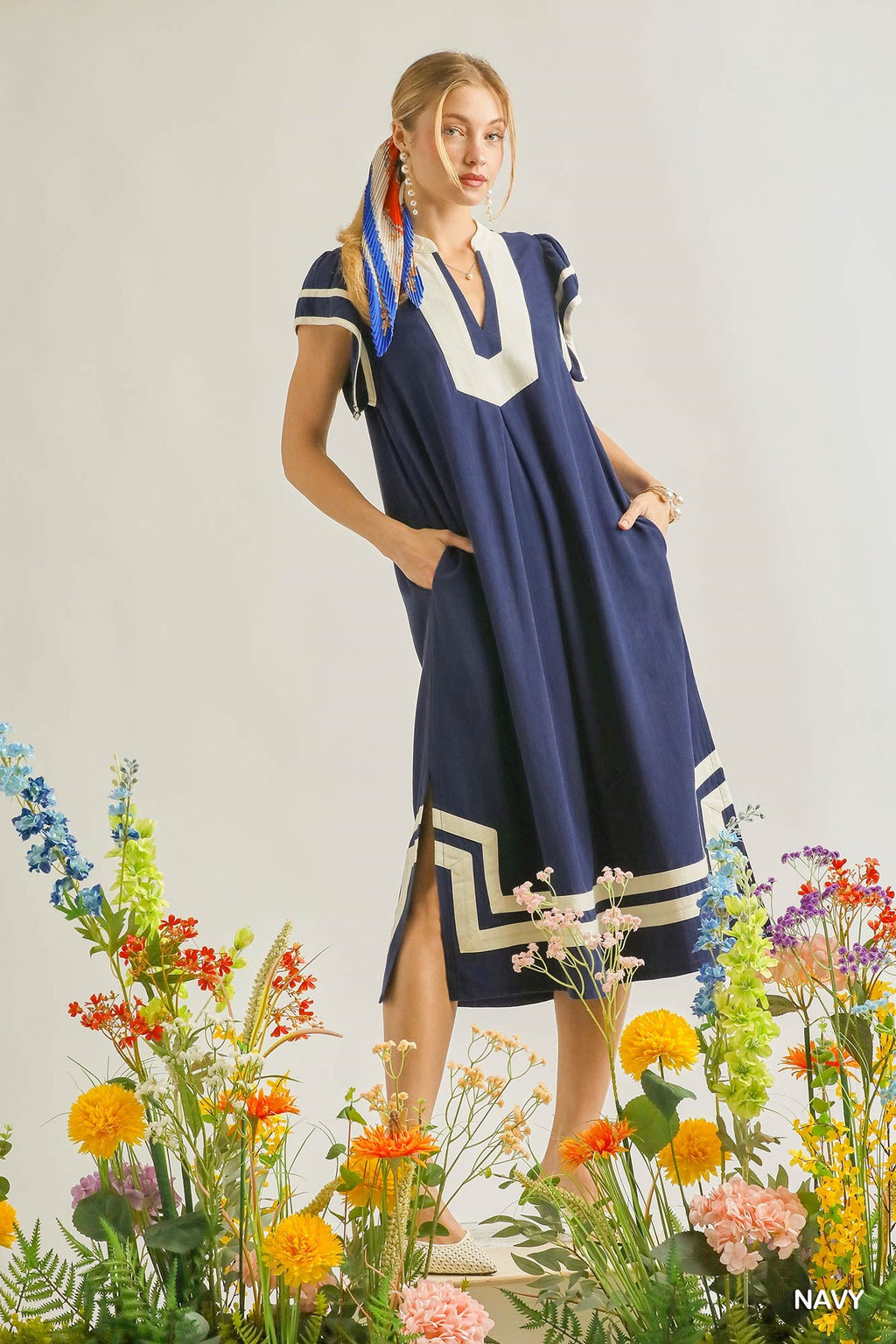Woman wearing a flowy navy midi dress with cream contrast trim, flutter sleeves, side pockets, and side slit detail in a relaxed resort-inspired silhouette standing among colorful flowers