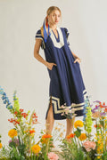 Woman wearing a flowy navy midi dress with cream contrast trim, flutter sleeves, side pockets, and side slit detail in a relaxed resort-inspired silhouette standing among colorful flowers