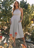 An outdoor full-length view of a model posing in front of large green cacti wearing a light sage green or grey textured halter midi dress. The dress features a gathered high-neck halter bodice, a wide fitted waistband, a flowy a-line midi skirt with side pockets, and is styled with strappy lace-up sandals