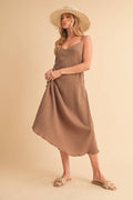 A woman posing in a sleeveless, solid brown crinkle gauze midi dress featuring thin adjustable cami straps, a subtle V-neckline, and an asymmetrical flowing hemline.