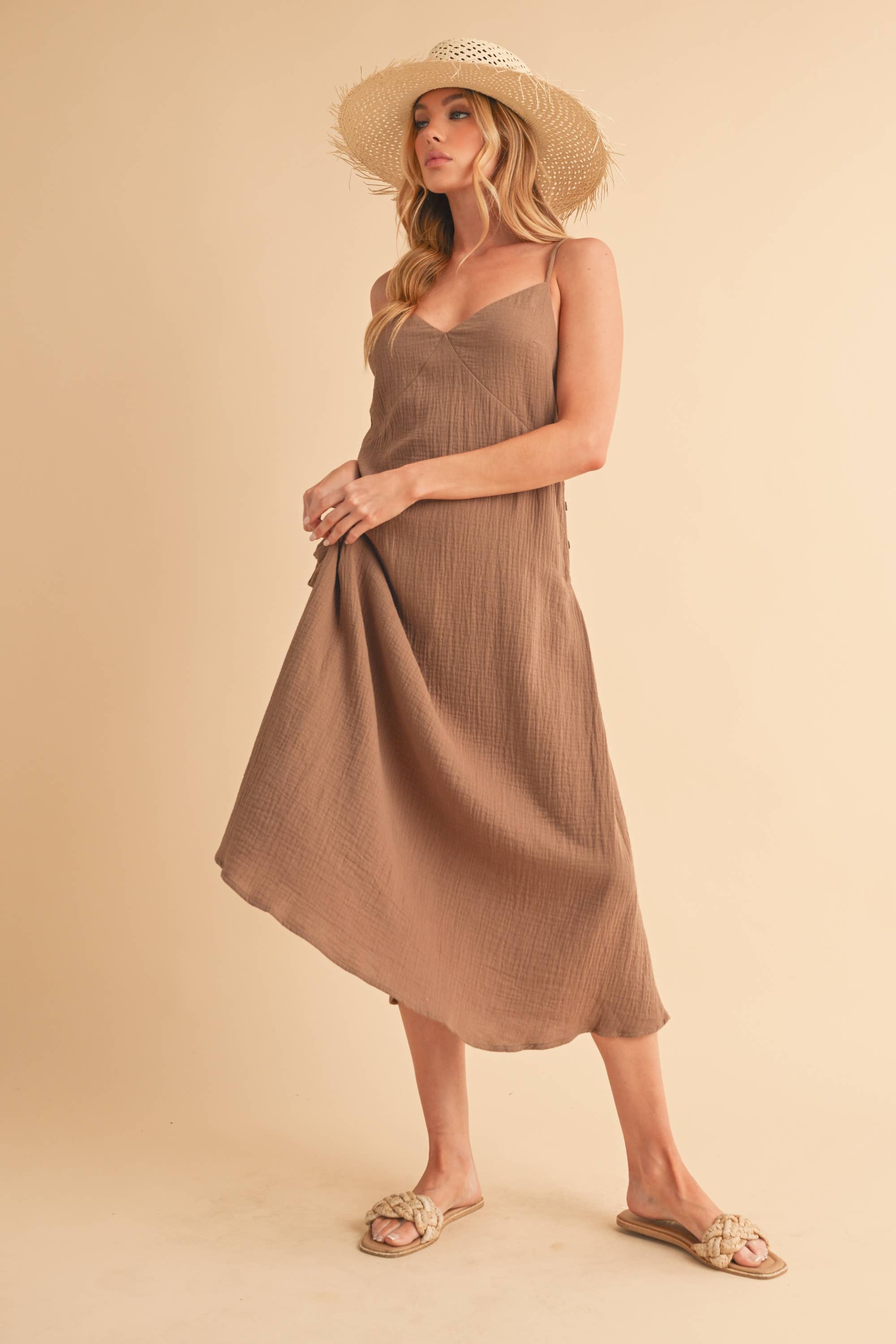 A woman posing in a sleeveless, solid brown crinkle gauze midi dress featuring thin adjustable cami straps, a subtle V-neckline, and an asymmetrical flowing hemline.