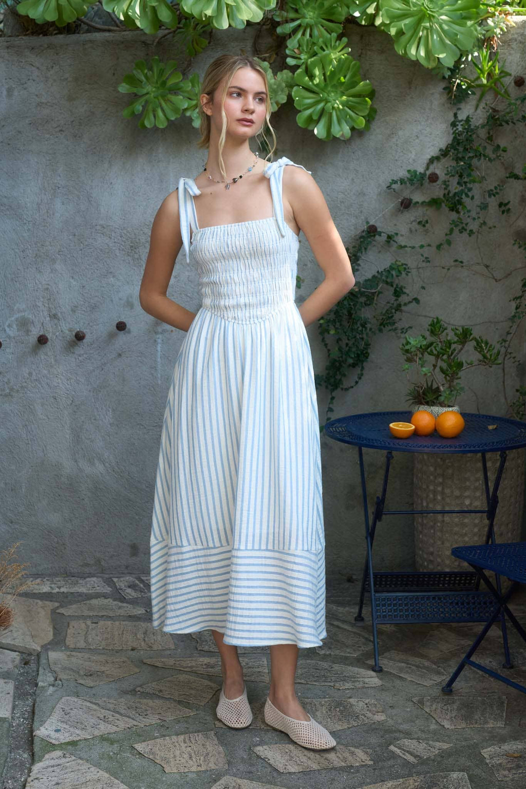 A woman posing outdoors wearing a white and light blue vertically striped midi dress, featuring a long smocked bodice, tie-strap shoulders, and a matching horizontal stripe pattern along the hemline standing outdoors with a stone wall and plants in the background.