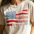 A close-up of a cream-colored t-shirt featuring a patchwork American flag graphic made of various fabric scraps including red gingham, blue florals, and white polka dots with exposed stitching.