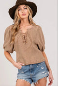 Woman wearing a tan waffle-knit lace-up top with puff sleeves and a chocolate brown wide-brim hat, paired with distressed denim shorts with  white background
