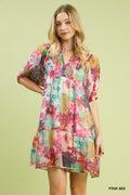 A woman wearing a colorful, abstract floral print mini dress with a v-neckline and smocked bodice detail. The dress features short puff sleeves, a tiered skirt, and hanging tassel ties. The print includes shades of pink, teal, yellow, and purple on a light cream background, set against a solid lime green backdrop.