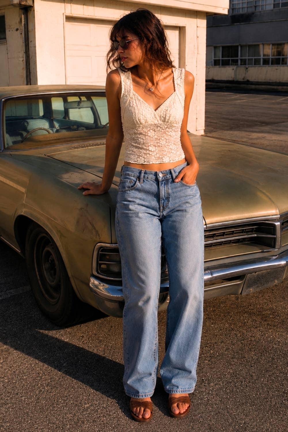 Woman in a white top and blue jeans standing next to a vintage car in an urban setting.